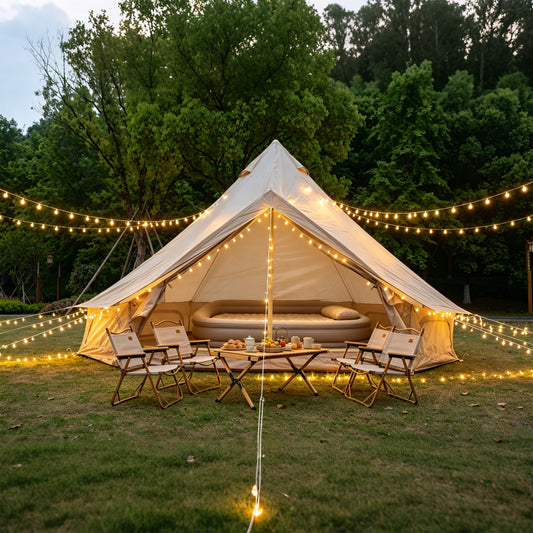 A large beige bell tent pitched on grass outdoors at dusk, surrounded by warm string lights and set with folding chairs and a small table. Trees fill the background.