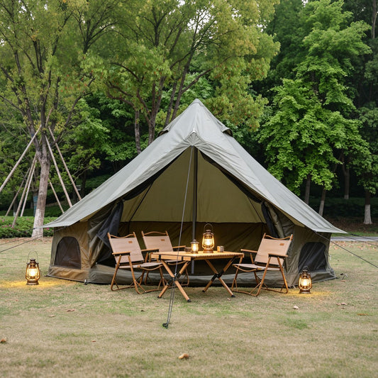 A large olive green bell tent pitched outdoors in a grassy clearing with trees behind it. A small camping table, four wooden chairs, and three lanterns are arranged inside the tent opening, creating a cozy campsite scene.