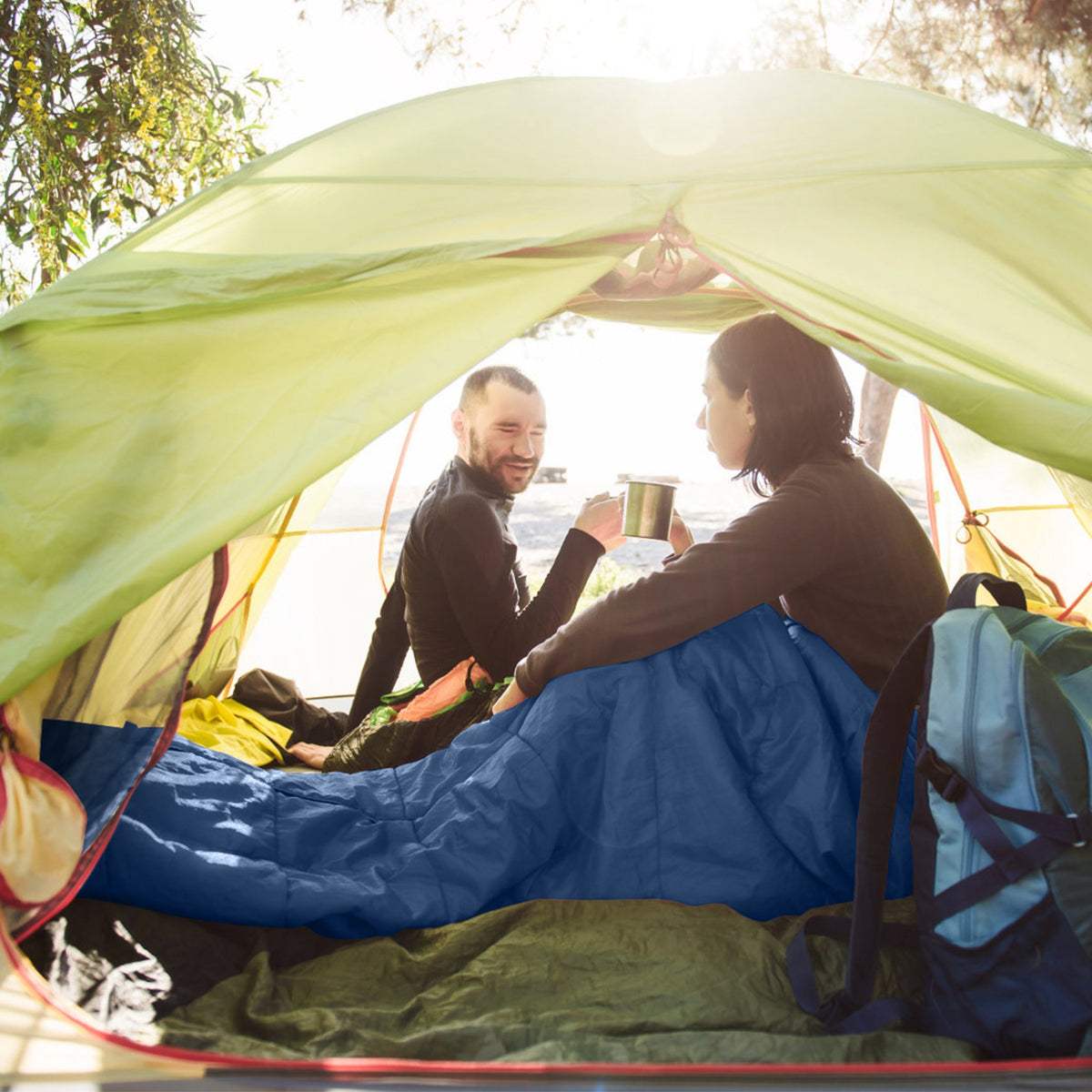 A couple sits inside a green camping tent sharing a mug, with a blue sleeping bag spread across the floor and a blue backpack in the foreground. Strong sunlight filters through the tent opening.