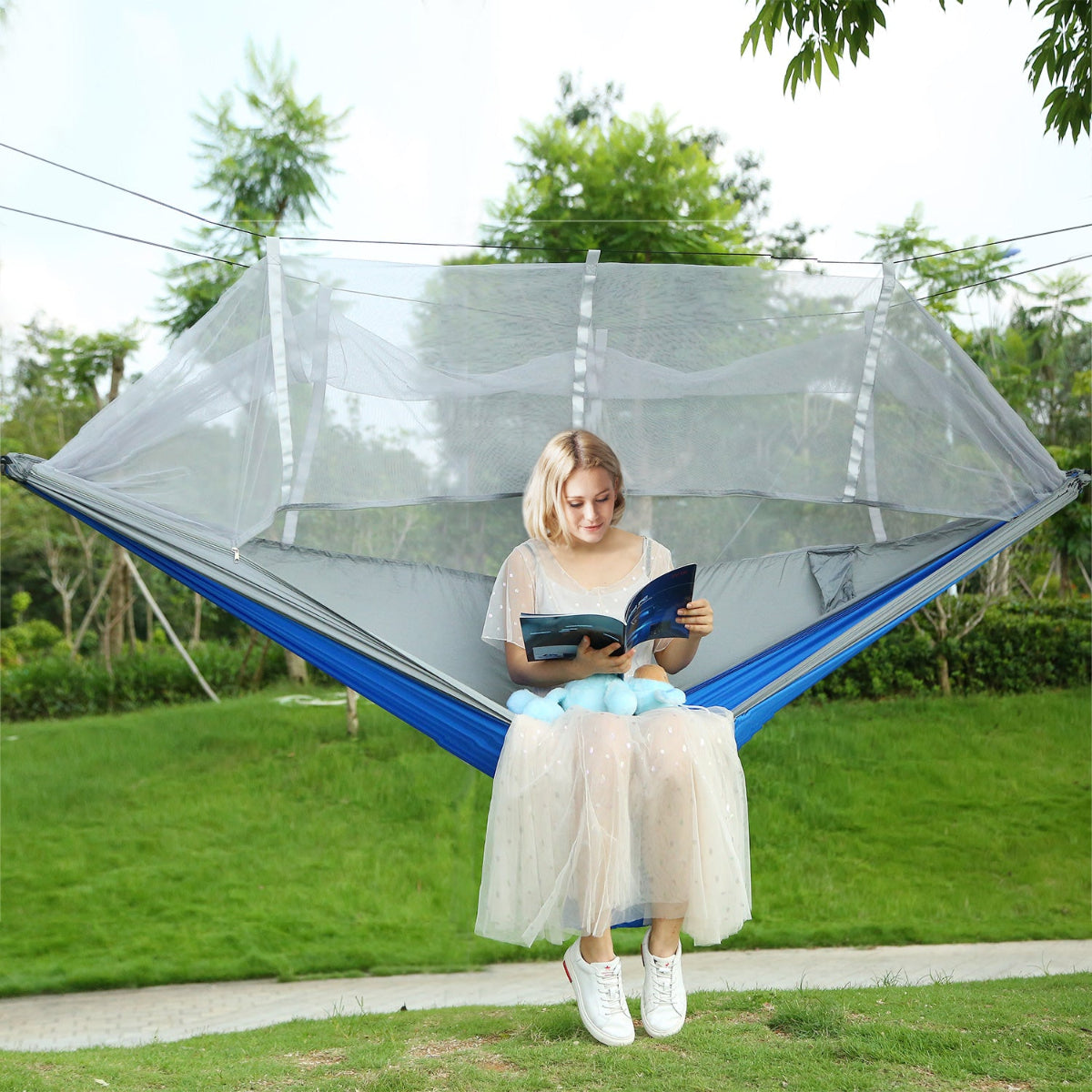 Woman reading a book in a blue hammock with mosquito netting outdoors.