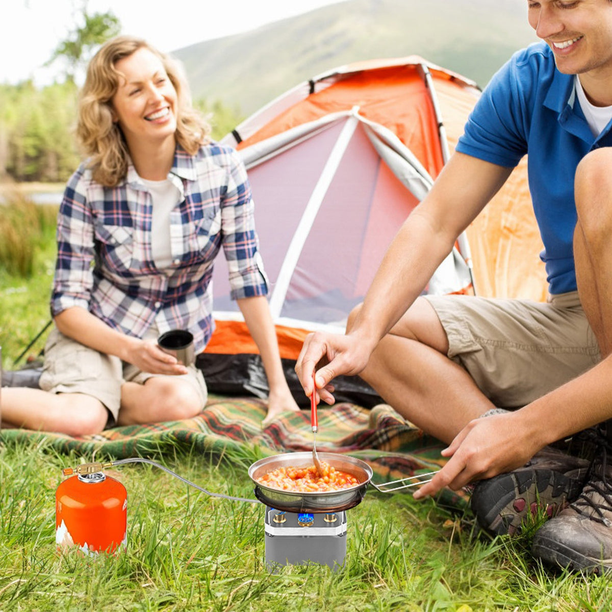 Two people camping outdoors, cooking on a portable stove with a tent in the background.