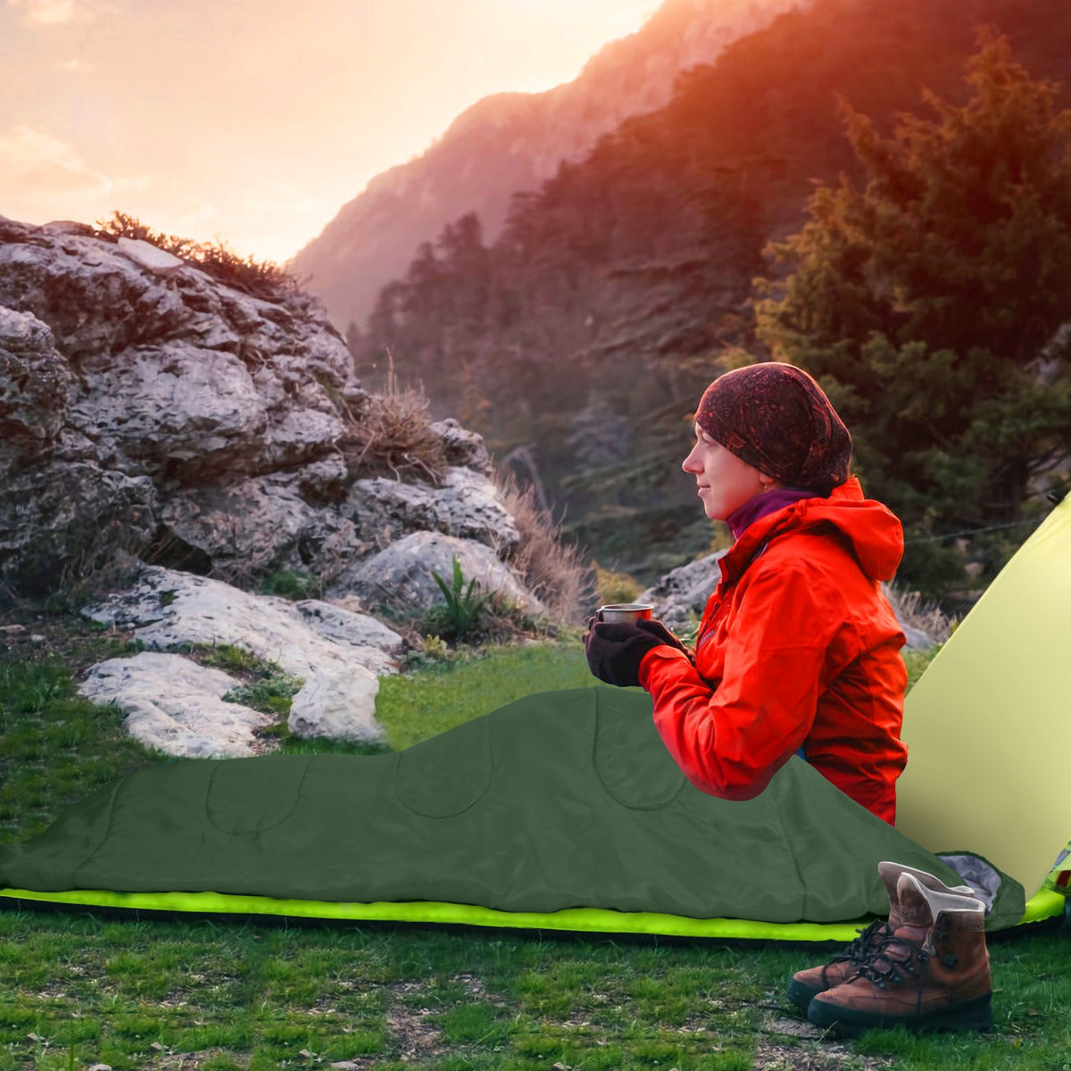 A person in a red jacket and brown beanie sits beside an olive green camping sleeping bag with a bright neon green trim in an outdoor mountain setting at sunset.