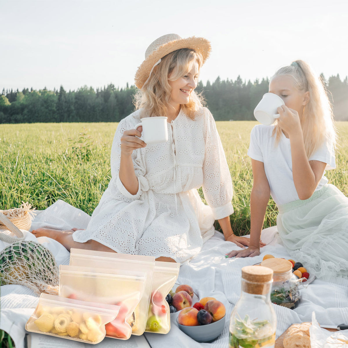 Two women sitting on a white picnic blanket in a grassy field, drinking from white mugs with fruit, bread, and clear storage bags in the foreground.