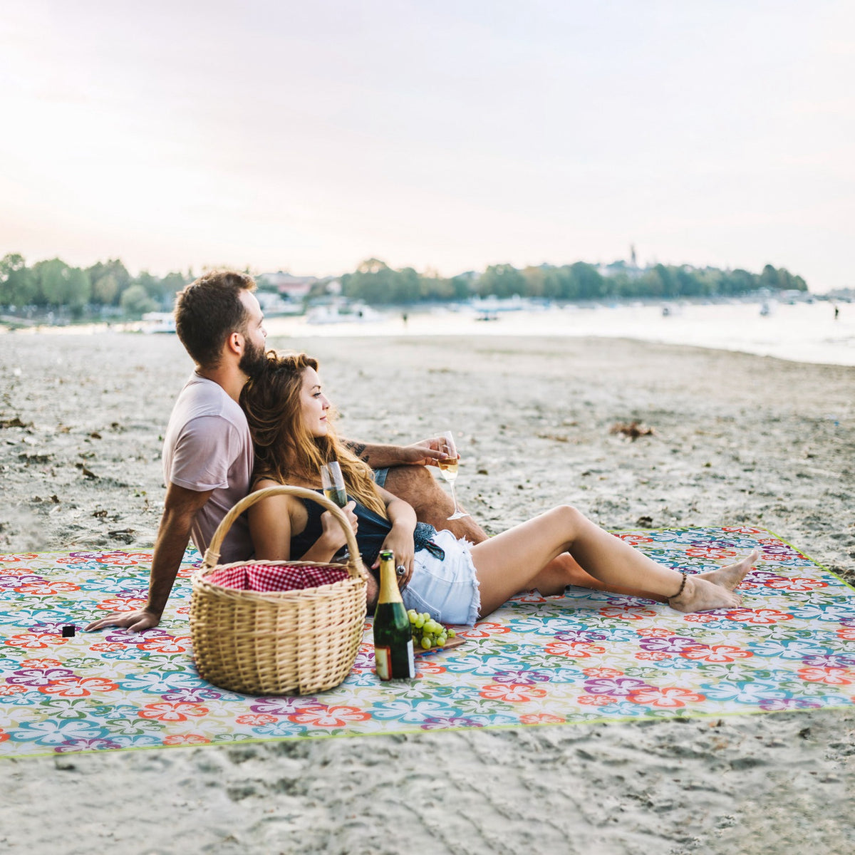 Two people sitting on a floral blanket by a beach with a picnic basket and bottle of wine.