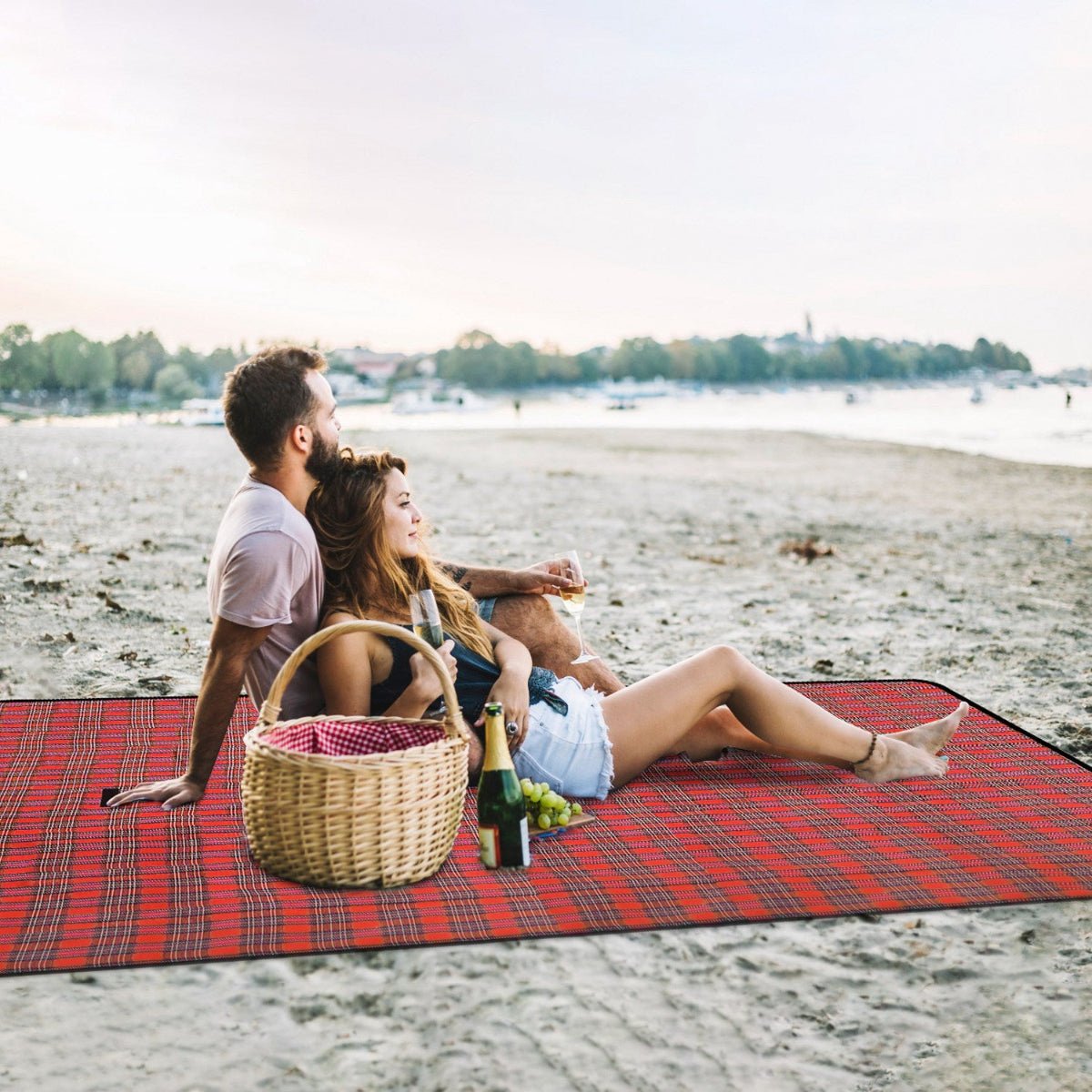 Red plaid picnic blanket spread on beach with two people sitting beside basket