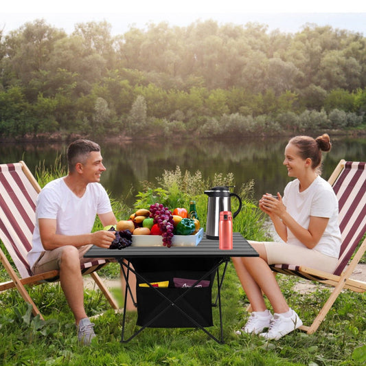 Portable aluminum camping table between two seated people beside a lakeside campsite