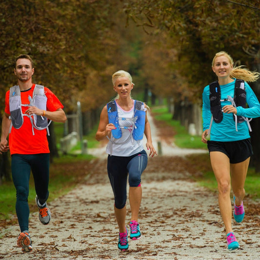 Three runners wearing hydration vests jogging on a tree-lined outdoor trail