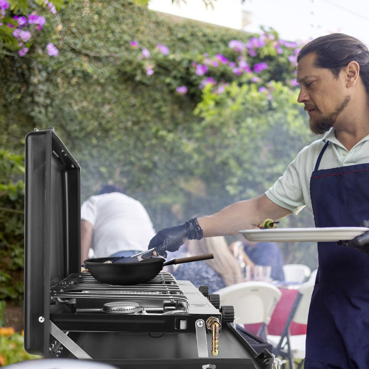 Person cooking on an open three-burner camping stove in an outdoor garden setting